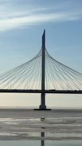 A low-angle shot presents a large, modern cable-stayed bridge set against a partly cloudy blue sky. The bridge features a tall, white, sharply-pointed central pylon from which numerous cables spread out to support the flat road surface. Cars are visible on the bridge. The bridge spans a flat, icy expanse of water, with reflections visible in the partially melted ice. The scene is still and quiet.