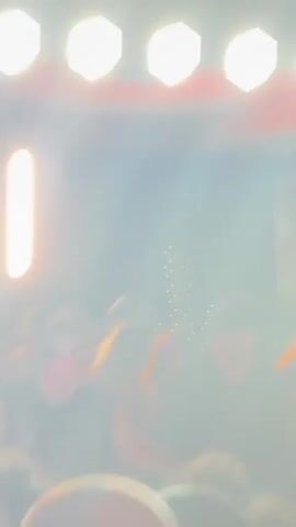 A video of two men DJing at a club or concert. The man on the left wears sunglasses and a black, sparkly vest. The man on the right wears a black shirt. They are standing behind DJ equipment, and a crowd of people is visible in front of them. The lighting is dim, with bright stage lights illuminating the performers and some of the crowd.