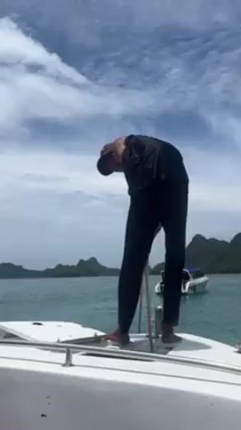 The video shows a man in a black sleeveless shirt and black pants standing on the bow of a white boat. He is pulling on a white rope attached to a metal pole on the boat. In the background, there is an expanse of blue water, islands covered with green trees, and another white boat. The sky is blue with scattered white clouds. The video appears to be taken on a sunny day.