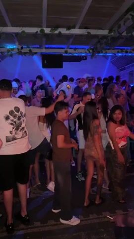 The video shows a group of young people dancing at a club. The lighting is dark, with blue and purple hues dominating the scene. Most of the dancers are casually dressed in summer attire, some wearing hats or caps. The music is loud and upbeat. There are green vines hanging from the ceiling. There is a speaker overhead. People are drinking and socializing. The atmosphere is energetic and festive.