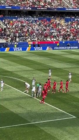 The video shows a soccer game from a high angle, focusing on a free kick situation. A group of players in white uniforms are lined up on one side of the ball, while players in red uniforms form a defensive wall. A referee in a green shirt is positioned between the two teams. The field is green with white lines, and the stadium seating is visible in the background, filled with spectators. Banners with "VISA" and "FIFA WORLD CUP 2026" are displayed.