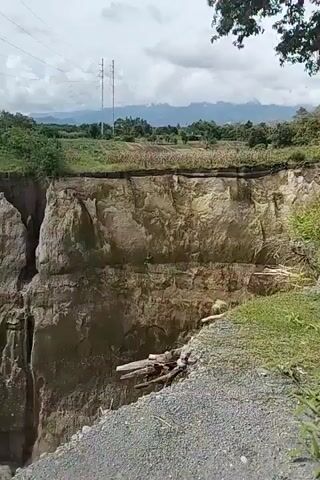 This media file shows a panoramic view of a dramatic geological formation, likely a canyon or ravine, in Kebunbaru, Indonesia. The camera pans across the landscape, revealing the sheer, eroded cliffs of the gorge. The walls of the canyon are layered with sediment, displaying various shades of tan, brown, and grey, with patches of green vegetation clinging to the upper edges and in sheltered crevices.

The scene is characterized by the stark contrast between the barren, weathered earth of the canyon and the lush green landscape that borders its rim. In the distance, rolling hills and mountains can be seen under a cloudy sky. Power lines stretch across the upper portion of the frame, hinting at nearby infrastructure. The ground in the foreground appears to be a gravelly path, suggesting the location is accessible, possibly a tourist spot or a natural landmark.

The activity taking place is the observation of this natural wonder, as indicated by the camera's movement and perspective. There are no people visible in the media file. The time of day appears to be daytime, with diffused light suggesting it might be overcast or that the sun is not directly overhead. The overall impression is one of awe at the power of natural erosion and the impressive scale of the landscape. The eroded surfaces and the dry riverbed at the bottom of the gorge suggest a history of water activity, perhaps flash floods, that have carved out this striking natural feature. There is no visible text in the media file.