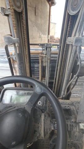 The video shows a first-person perspective from inside a dirty forklift. A hand is visible on the steering wheel. The mast and chains of the forklift are in the foreground, with a stack of wrapped objects visible in the background. The environment outside the forklift appears to be a construction or storage yard. There is writing on the dashboard. The hand is visible adjusting the steering wheel.