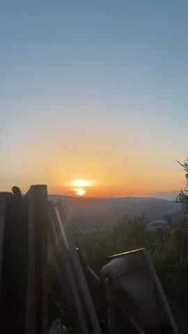 The video shows a sunset over a rural landscape in Svinjišta, Montenegro. The camera is positioned on a hill, overlooking a valley with a small town and mountains in the distance. The sun is setting on the horizon, casting a warm glow over the scene. The sky is clear and blue, with a few wispy clouds. Trees and bushes are in the foreground, obscuring some of the view. There is also a pile of debris in the foreground, including plastic bags and what appear to be broken pieces of wood or metal. The overall mood of the video is peaceful and serene, with the beauty of nature taking center stage. The time of day is clearly sunset. There is no visible text in the video.