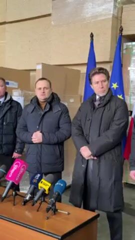 The video shows three men standing behind a table with microphones in front of them. The two men in the center are the focus, both wearing dark winter coats. Behind them are stacks of boxes and two flags with gold stars on a blue background. The man on the left is speaking and gesturing with his hands. The man on the right has his hands clasped and then briefly adjusts his hair.