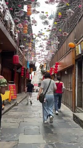 People walk down a narrow street paved with stone. Overhead, a net is decorated with various colorful flowers and orange spheres. Red lanterns hang along both sides of the street, which is lined with old wooden buildings.