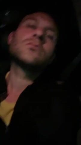 A man is in a car at night. He is wearing a black jacket over a yellow shirt. He has short, curly brown hair and a beard. The lighting is dim, with a red and yellow hue. His expression is serious. He appears to be looking at the camera and then away. The video is shot from a low angle.