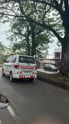 The video shows a street scene from the perspective of a vehicle, likely a motorcycle, based on the visible mirror on the left side of the frame. On the left side of the road, an ambulance with red and white markings and a red cross symbol is driving away from the camera. Further down the road on the left, there are several parked vehicles and a sidewalk lined with trees and some low buildings. On the right side of the road, there are more trees, some buildings with corrugated metal roofs, and more parked vehicles, including what appears to be a black pickup truck. The sky is overcast, suggesting a cloudy day. The road is paved and has a white line marking the edge. The overall scene appears to be in a developed area, possibly a town or city.