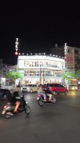 At night, traffic weaves through a busy intersection. The camera captures a street scene dominated by a brightly lit, two-story building with the word "TOKY" displayed in neon on its roof. Cars and scooters zip through the intersection, headlights cutting through the darkness. The building appears to be a retail store, judging by the displays visible through its large windows. Pedestrians can be spotted inside the store as well. Trees flank the building, adding a touch of greenery to the urban setting.