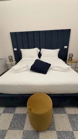 The video shows a bedroom. A king-sized bed with a dark blue headboard and frame is centered in the frame, covered in white linens and topped with folded white towels and a dark blue pillow. A golden yellow velvet stool sits in front of the bed on a gray and white checkered tile floor. Two small round side tables are located on either side of the bed, each with a small lamp. The walls are white, and a decorative metal piece hangs on the wall to the left. An air conditioning unit is visible below the decoration. The camera pans upward to show an ornately decorated ceiling with painted murals and designs in muted blues and browns.