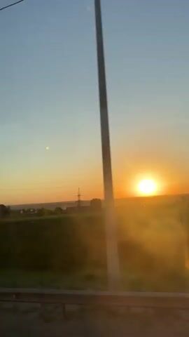 The video shows a bright sunset over a rural area, viewed from a moving vehicle. The sun, a bright orange orb, sits on the horizon, with light rays streaming outwards. Silhouetted against the sunset are various structures including a water tower and power lines. In the foreground, there is a guardrail, indicating the view is from inside a vehicle moving along a road. The sky transitions from a light blue at the top to orange near the horizon. The environment seems to be a mix of fields and scattered houses.