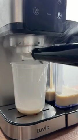 A close-up shot shows a coffee machine dispensing liquid into a clear plastic cup. The cup already contains a small amount of light brown liquid. The machine is silver and black, with buttons and indicators visible on its front panel. Steam is rising from the machine as it dispenses liquid. To the right, two clear containers are partially filled with a similar light brown liquid. The cup sits on a silver, slatted tray. A black spout from the machine is positioned directly above the cup.