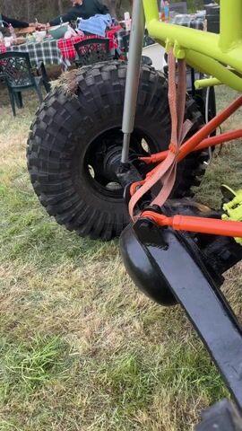 The video shows a custom-built off-road vehicle, likely a buggy or modified ATV. The vehicle has a tubular frame painted a bright yellow-green. Orange suspension components are visible, and FOX-branded shocks are mounted on the frame. The tires are large and knobby, designed for off-road traction. The engine and drivetrain are exposed, suggesting a minimalist design focused on performance. The vehicle is parked on a grassy area, with some household items and furniture visible in the background. The lighting suggests it is either early morning or late afternoon. The camera moves around the vehicle, showing different angles and details.