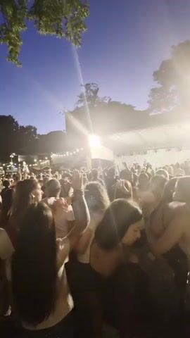 The video shows a crowd of people at an outdoor event, possibly a concert, at dusk. The sky is a deep blue, with some trees visible at the top of the frame. A stage with bright lights and possibly performers can be seen in the background, with a large group of people gathered in front of it. The crowd appears to be diverse in age and gender, with many people facing the stage and some with their hands raised. The lighting is dim, with spotlights from the stage illuminating parts of the crowd. The overall atmosphere seems energetic and festive.