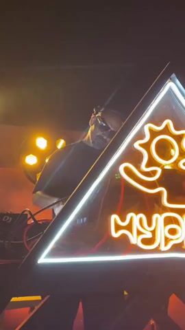 The video shows a person wearing sunglasses and a dark t-shirt behind a DJ setup with neon lights and stage lighting. The person appears to be a DJ performing at a club or event. The background is dark with colorful stage lights. The video also shows a blurry crowd of people in the background, suggesting a lively atmosphere.