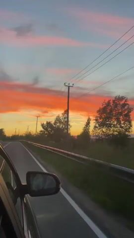 The video is shot from inside a car looking out the passenger window as the car drives along a paved road. The road has a white line dividing the lanes. A metal guardrail runs along the side of the road. Trees and vegetation line the side of the road. In the background, there are telephone poles and power lines. The sky is a vibrant sunset with streaks of orange, pink, and purple clouds. The sun appears to be setting behind the trees on the horizon. The overall scene depicts a scenic drive during sunset.