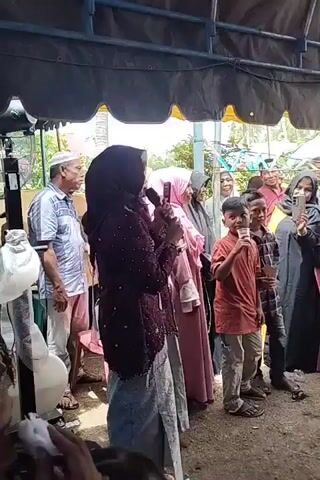 A group of people are gathered outdoors under a tent. A woman in a dark headscarf and embroidered top is holding a microphone and a cell phone, speaking. To her right, several other women are also wearing headscarves, and one is looking at her phone. A boy in an orange shirt is to the right of the women, holding a cup. To the left of the woman speaking, a man wearing a white cap and a patterned shirt is standing. A child's toy is partially visible in the foreground.