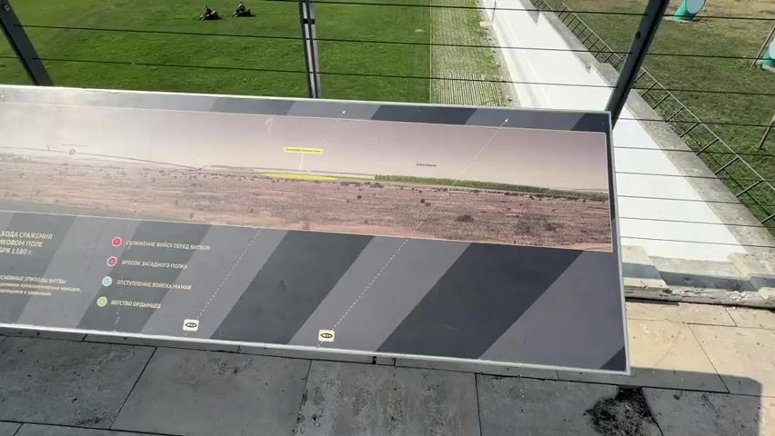 The video shows a long, wide, horizontal interpretive panel with a panoramic image of a battlefield. The panel has a black and gray striped border at the bottom. Text and graphics are overlaid on the panoramic image, depicting troop movements and key locations. A legend with color-coded markers and corresponding descriptions is visible on the lower left of the panel. The panel is positioned outdoors, likely at a historical site. In the background, a grassy area with two people sitting can be seen, as well as a white-tiled walkway and a fence. The lighting suggests it is daytime. The camera is positioned at an angle looking down at the panel.