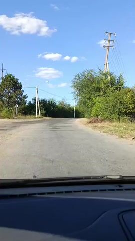 The video shows a first-person view from inside a vehicle driving on a paved road. The road is in fair condition with some potholes and cracks. The vehicle is moving forward on a slight uphill incline. On the left side of the road, there are trees and utility poles with wires. On the right side of the road, there are trees and grassy areas. The sky is blue with scattered white clouds, indicating a daytime scene. There are two traffic signs visible on the right side of the road, one appears to be a circular sign with a red border and a horizontal line, likely indicating "no entry" or a similar restriction, and another sign above it which is less clear but may be related to speed limits. The overall environment suggests a rural or suburban setting on a clear day.