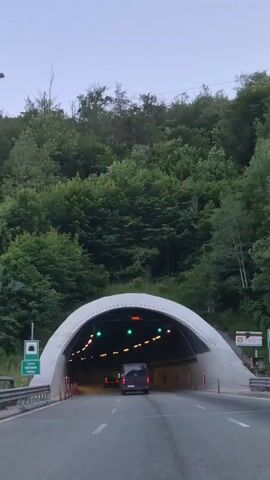 The video shows the interior of a road tunnel. The perspective is from inside a vehicle, suggesting the video was filmed from the driver's or passenger's seat. The tunnel is illuminated by rows of yellow lights along the ceiling and green directional arrows are visible overhead. Orange signs are also present. The road is paved and marked with white lines. Several other vehicles, including a dark-colored van and two red cars, are visible further ahead in the tunnel. The tunnel walls are concrete, and there are orange traffic cones and red and white safety markers along the sides. The entrance to the tunnel is visible at the beginning of the clip, showing a curved white structure and green trees surrounding the opening.