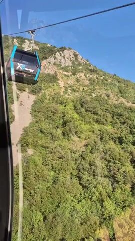 The video shows a cable car moving along a cable line against a backdrop of a lush green mountainside. The cable car is blue and black with the logo "Xenergy" on its side. The camera angle appears to be from inside another cable car, looking out. The sky is clear and blue. The video captures the ascent of the cable car over a forested area, with glimpses of the landscape below, including water bodies and distant structures. Sunlight streams in from the side, creating lens flare effects.