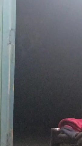 The video shows a dark room with a light blue doorframe visible on the left side of the frame. The room appears to be dimly lit. Towards the bottom right of the frame, there is a folded red cloth or blanket on top of a dark object which is possibly a chair or container. The lighting is inconsistent and casts long shadows. There is some audio present with someone speaking in the background.