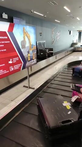 The video shows luggage arriving on a baggage carousel in an airport. Several suitcases of different colors and sizes move along the black conveyor belt. In the background, people are waiting to claim their luggage. One side of the carousel is lined with advertising posters. The airport environment appears modern and well-lit.