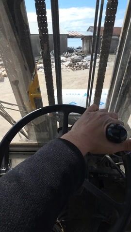 The video shows a first-person view from the inside of a forklift. A hand in a black long-sleeved shirt is holding the steering wheel. The forklift is stationary. Outside the forklift, through the cage and window, is a yard with a building and debris scattered around. There are also stacked building materials visible. The sky is blue and visible in the background. The forklift interior is dusty and appears well-used.