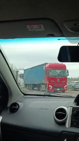 The video is shot from the inside of a parked car. From the point of view of the driver, a red SHACMAN X5000 truck is blocking the road ahead. A white truck is to the left of the SHACMAN. A silver car is also parked behind the red truck. The sky is overcast. The interior of the car is visible in the foreground. The radio is visible in the dashboard. Music can be heard.