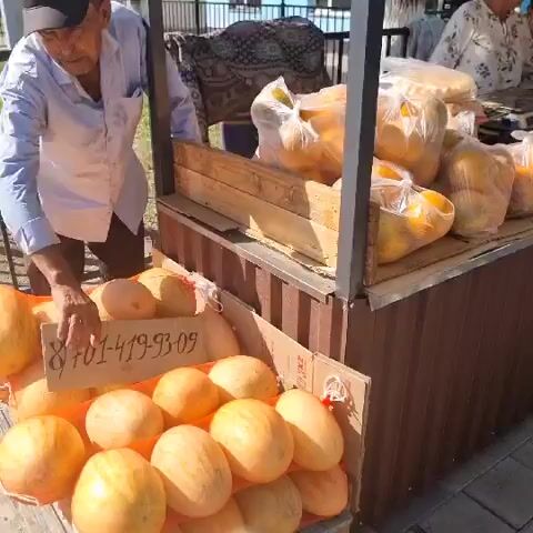 The video shows an outdoor market stall selling fruits and bread. An older woman in a headscarf and patterned shirt is serving a younger person. There are bags of oranges, melons, peaches, cherries, and flatbreads on display. A scale is visible on the counter. The stall is made of wood and metal, and there is a fence in the background. The lighting suggests it is daytime.