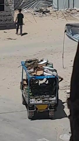 The video shows a view of a refugee camp. In the foreground, a three-wheeled vehicle is loaded with boxes and bags. In the middle ground, a person is walking away from the camera. There are several makeshift stalls with signs written in Arabic. Some of the stalls appear to be selling food, as indicated by images of ice cream cones and the word "murbat" which means jam or preserves. The background shows rows of tents and temporary structures, characteristic of a refugee settlement. The overall environment appears dusty and weathered.