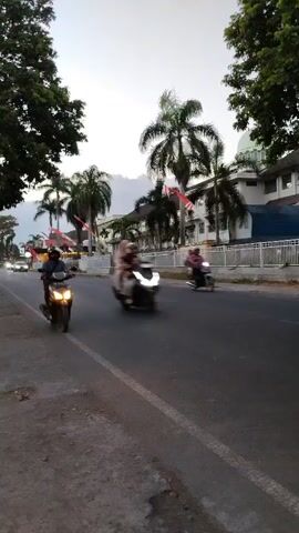 The video shows a street scene at dusk. A motorcycle rider wearing a helmet is moving away from the camera on the left side of the road. In the background, there are palm trees and a building with a dome, possibly a mosque or a government building, adorned with Indonesian flags. Cars are also visible on the road, one of which is in motion and appears blurred due to the camera's movement. The lighting suggests it is either dawn or dusk.