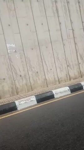 The video captures a view from a moving vehicle, possibly a tricycle, traveling alongside a large concrete wall. The wall appears to be part of an overpass or elevated structure, with vertical lines and some minor staining visible. A sidewalk with alternating black and white painted curbs runs between the wall and the road. The road surface is asphalt. In the foreground, a portion of the vehicle's frame, likely the side of the tricycle, is visible, painted yellow and black. Further down the road, the tricycle itself, with a black canopy, is briefly visible. The video then pans to show the underside of the overpass structure, with its concrete pillars. Beyond the overpass, a brighter, open area is visible with trees, utility poles, and what appear to be several parked tricycles and a few people gathered near a market-like setting.