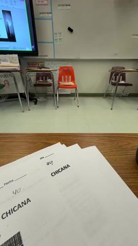The video shows a classroom setting. In the foreground, there are sheets of paper with the word "CHICANA" printed on them along with the number "40". Beyond the desk with the papers, there are several student desks and chairs arranged in the room. The chairs are of different colors, including red and maroon. In the background, there's a whiteboard with some writing and images on it. A screen is also visible, displaying a digital image. A person is seen standing near the screen. The camera seems to be positioned on a desk, capturing the classroom scene. The room appears to be well-lit and organized.