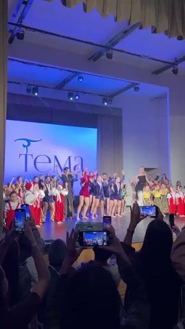The video shows a stage performance by a large group of children, likely a dance recital. The stage has a large screen in the background with the logo "Tema" and some smaller text beneath it. There are approximately 30 children on stage, split into groups with different costumes. One group wears red and white traditional outfits, another wears red velvet outfits, and a third wears navy blue outfits. Three adults, two women and one man, stand among them, presumably instructors. The audience is visible in the foreground, holding up phones to record the performance. The room is lit with blue and white stage lights. The overall atmosphere is festive and focused on showcasing the children's performance.