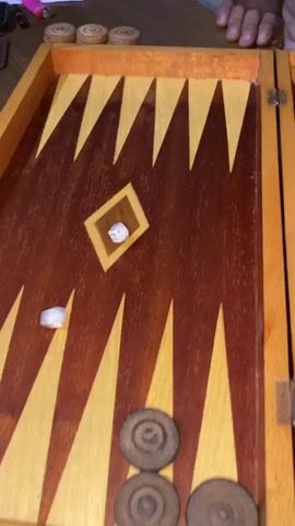 A close-up, overhead shot of a wooden backgammon board with dark red and light wood triangular patterns. Two white dice are visible on the board, one showing a 6 and the other showing a 3. One die is near the center of the board, above a diamond-shaped inlay, and the other is lower down, closer to the viewer. The board is set up for a game, and the pieces are not visible. The lighting is dim.