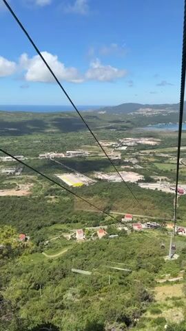 A scenic daytime video captures a view from a cable car ascending or descending over a lush green landscape. The camera looks out over a vast valley dotted with buildings, dense trees, and a large flat area, possibly an industrial site or airport, in the middle distance. In the far background, the blue sea meets the horizon under a bright sky with scattered white clouds, with distant mountains or hills lining the coastline.

Cable car lines stretch across the frame. At 0:02, another cable car, dark in color with windows, approaches and passes by, heading in the opposite direction. The word "Priceless" is visible on the side of the passing cable car. The view indicates a significant elevation, offering a panoramic outlook over Čavori, Montenegro, towards the Adriatic Sea. The weather is clear and sunny.