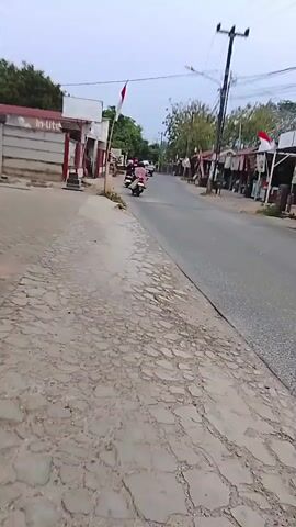 A person is filming the ground and a paved road with some shops on the right. There are a few motorcycles on the road, and some Indonesian flags are visible. The sky is overcast, and it appears to be either early morning or late afternoon.
