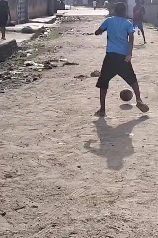 The video shows a group of boys playing soccer on a dirt road in what appears to be a residential area. They are kicking a ball around, trying to score or pass to each other. There are buildings in the background, and some litter is scattered along the roadside. The lighting suggests it's daytime.