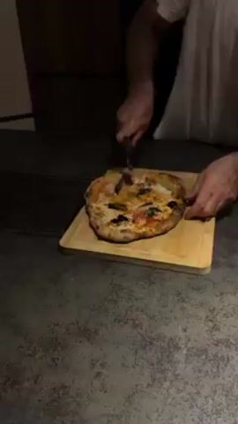 A pizza is being cut with a pizza cutter on a wooden board. The pizza has a charred crust and is topped with cheese, tomato sauce, basil, olives, and what appears to be sliced green peppers. The person cutting the pizza is wearing a white t-shirt and using a rolling pizza cutter. The pizza is being cut into six slices. The surface the pizza is on is a dark, mottled grey. The light is dim.
