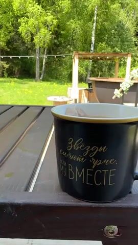 A close-up shot shows a black mug sitting on a brown wooden picnic table, outdoors. The mug is filled with liquid and has a teabag inside. Gold lettering on the mug reads "Звёзды сияют ярче, если мы вместе", which translates to "Stars shine brighter when we are together". The background features a green lawn with scattered yellow flowers, various trees, and a wooden structure with a hot tub-like feature. There is a light string hanging in the background. The light is bright and sunny.