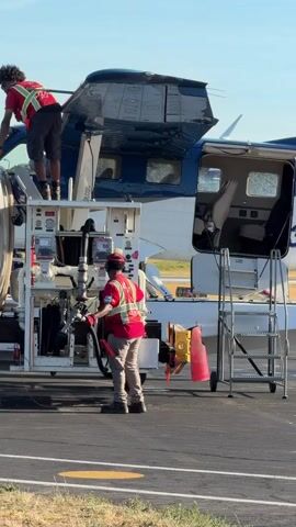 Two individuals in high-visibility vests are refueling a small aircraft on an asphalt surface, likely an airport tarmac. The person on the left is standing on top of a refueling truck, leaning towards the aircraft's wing. The person on the right is standing on the ground next to the refueling truck, holding a fuel hose. The aircraft is blue and white, with its passenger doors open. A set of stairs is positioned next to the aircraft's entrance. The scene is illuminated by bright sunlight, casting shadows.