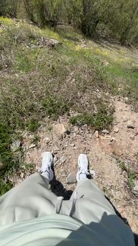 From a first-person perspective, the video pans up from a person’s feet standing on a rocky, grassy hillside to reveal a vista of rolling green hills. The person is wearing light gray sweatpants and white sneakers. The ground beneath their feet is covered in a mixture of rocks, dirt, dried grass, and some green grass. As the video pans up, the view opens to a landscape of densely forested hills under a bright blue sky. A landslide scar is visible on one of the hillsides.