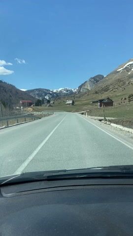 The video shows a view from inside a car driving on a paved road in a mountainous area on a clear, sunny day. The road has a single white line down the center. In the distance, snow-capped mountains are visible. There are several small buildings or houses scattered along the roadside, some with wooden construction. The landscape consists of grassy fields and some trees. The car's dashboard is visible at the bottom of the frame. There is minimal movement within the frame besides the forward motion of the car.
