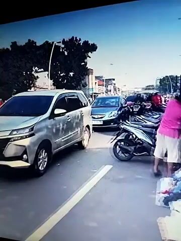 The video shows a street scene with several vehicles and people. In the foreground, a person wearing a blue shirt and a backpack is walking away from the camera. A silver car is parked on the right side of the road. In the background, there are buildings, trees, and a blue van. A person on a red motorcycle is also visible. The scene appears to be an urban environment. The timestamp at the beginning of the video is 00:00.