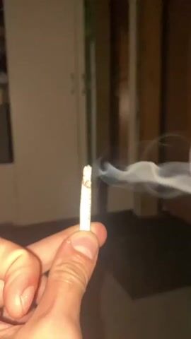 The video shows a close-up of a hand holding a lit cigarette from which smoke is rising. The background is out of focus and appears to be an indoor environment with dim lighting. The smoke forms swirling patterns as it drifts away from the cigarette.