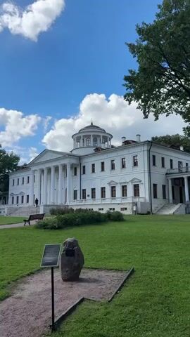 The video pans across a manicured park in front of a grand white neoclassical building with a dome. The building features a prominent colonnade. In the foreground, a large rock with a plaque is situated on a gravel path. Benches are placed on the lush green lawn. Several people are scattered throughout the park, some walking, some sitting on benches, and a few are congregated near the building entrance. The sky is blue with scattered white clouds, and trees line the perimeter of the park.