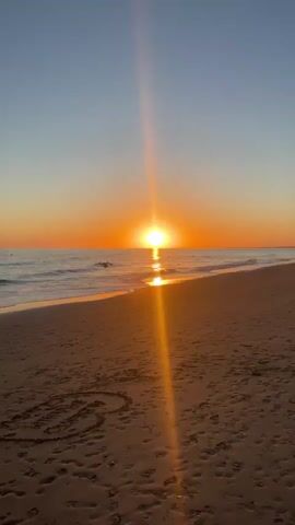 The video shows a beach at sunset. The sun is setting over the ocean, casting a warm orange glow on the sky and water. The sand is wet from the tide, and there are footprints in the sand. In the distance, there are buildings and people walking on the beach. The video also shows a couple walking on the beach.