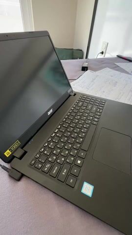 The video shows a black Acer laptop resting on a bed with a light purple or gray bedspread. The laptop is open, and the screen is dark. The keyboard has both English and Cyrillic characters. To the right of the laptop, there are several sheets of paper covered in handwritten notes, along with a pen and a small black device. In the background, there is a window to the left and a whiteboard with a dark frame on the right, partially obscured by what appears to be a piece of artwork or a drawing. There is also a green bag or soft case visible behind the laptop. The overall impression is of a workspace set up on a bed. The speaker says "Work, work, go to Fedota."