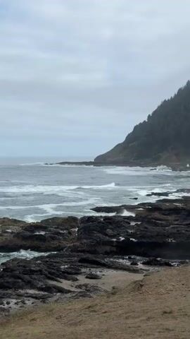 The video shows a rocky coastline with waves crashing against the shore. A forested hill rises steeply from the water. The sky is overcast, and fog is visible in the distance, obscuring the top of the hill. The foreground consists of a dirt path and sparse vegetation. The waves are white and foamy, indicating rough conditions. The rocks are dark and jagged, appearing wet from the sea spray. The overall atmosphere is somber and natural. No people or animals are visible.